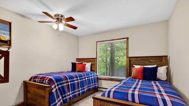 Quarto com duas camas de madeira, um ventilador de teto, uma janela com persianas e um quadro na parede.
