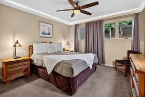 Un dormitorio con cama, mesitas de noche, escritorio, silla, ventilador de techo y ventana con cortinas.