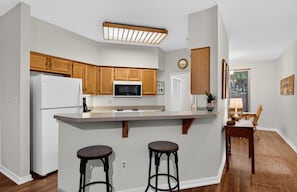 A kitchen with wooden cabinets, a white refrigerator, a microwave, and a bar area with stools. - Trillium Close 18 with Wintergreen Advantage (Wintergreen)