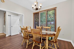 A dining area with a round table and six wooden chairs, a chandelier, and a window with blinds. - Trillium Close 18 with Wintergreen Advantage (Wintergreen)