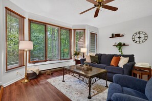 A living room with a ceiling fan, large windows with blinds, a sofa, a coffee table, and a wall clock. - Trillium Close 18 with Wintergreen Advantage (Wintergreen)