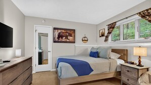 Ein Schlafzimmer mit Bett, Nachttischen, einem Schminktisch, einem Fernseher und einem Fenster mit Jalousien.