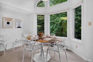 Ein Essbereich mit einem runden Tisch, gedeckt für vier Personen, umgeben von weißen Stühlen, großen Fenstern mit Blick auf Grünflächen und zwei gerahmten Kunstwerken an der Wand.
