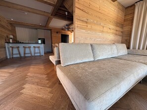 A modern living room with a sofa, wooden flooring, and a kitchen in the background. - New Chalets in the Tannheimer Valley (Jungholz)