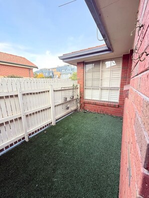 A fenced backyard with artificial grass, a brick wall, and a window with shutters.