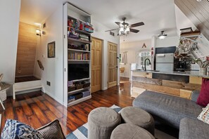 A modern living room with a television, bookshelves, a sofa, and a kitchen area with a refrigerator and a sink.