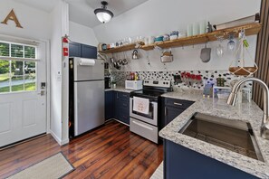 A modern kitchen with a refrigerator, stove, oven, and microwave. There is a wooden floor, a window with blinds, and a door.