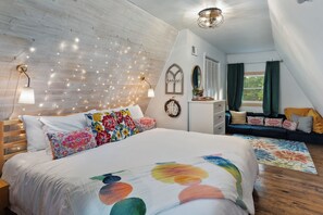 Une chambre avec un plafond en bois, un lit recouvert d’une literie à motifs floraux colorés, une table de chevet, une commode, une fenêtre avec des rideaux et un luminaire fixé au mur.