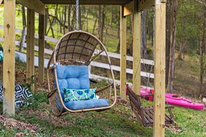A wicker swing chair with blue cushions and a patterned pillow, suspended from a wooden structure with a white fence in the background.