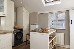 A compact kitchen with a built-in washing machine, a small dining area with a table, and a window with blinds. - Thomas Street House -  a cottage that sleeps 6 guests  in 3 bedrooms (Cirencester)