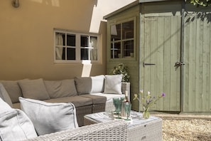 A wicker sofa with cushions, a table with a bottle and glasses, and a green wooden door. - Thomas Street House -  a cottage that sleeps 6 guests  in 3 bedrooms (Cirencester)