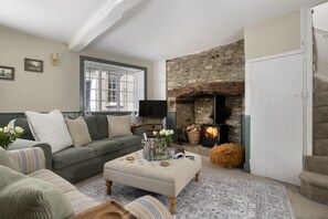 A cozy living room with a stone fireplace, a sofa, a television, and a decorative ottoman. - Thomas Street House -  a cottage that sleeps 6 guests  in 3 bedrooms (Cirencester)