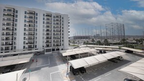 Een meerlaags woongebouw met balkons, een parkeerplaats met overdekte parkeerplaatsen, en een groot stadion op de achtergrond.