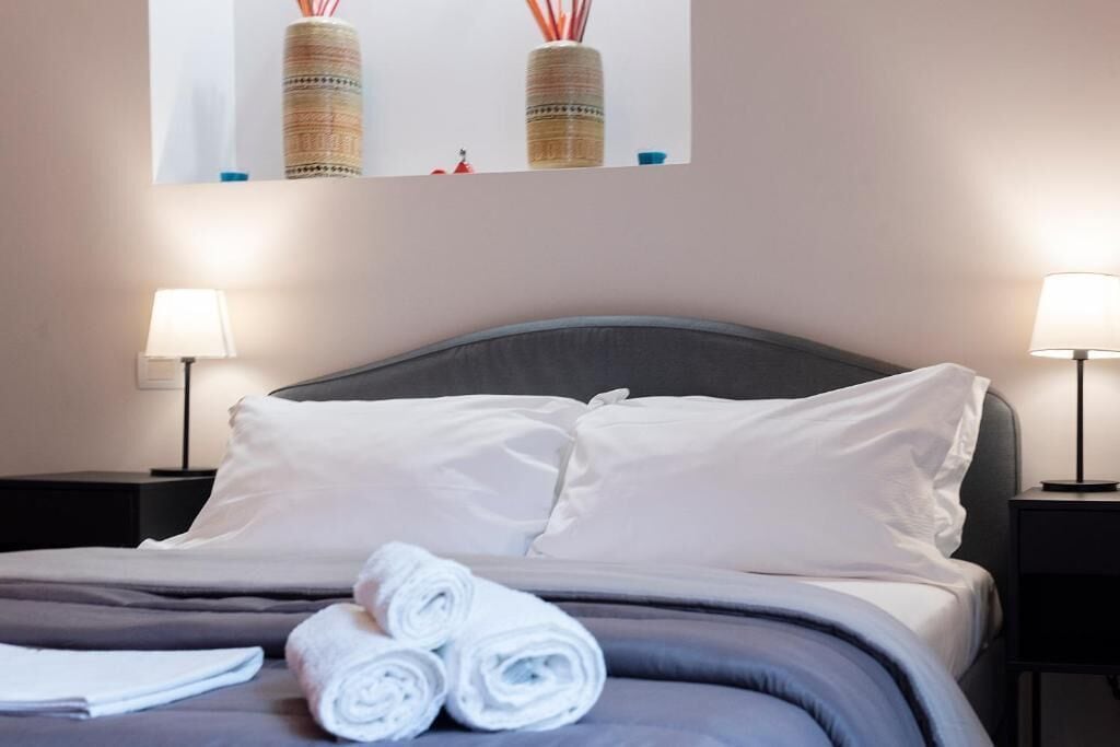 Ein ordentlich bezogenes Bett mit weißen Leinen und aufgerollten Handtüchern, zwei Nachttischlampen und ein gerahmtes Bild über dem Bett.