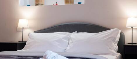 Una cama bien hecha con sábanas blancas y toallas enrolladas, dos lámparas de noche y un cuadro enmarcado sobre la cama.