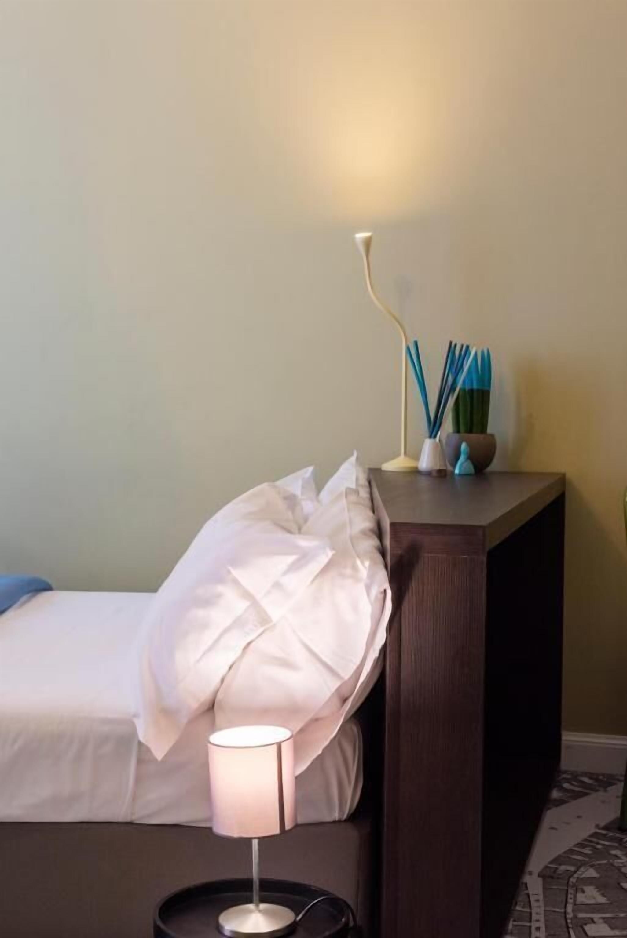 A hotel room with a bed, a bedside lamp, a nightstand with a lamp and a small plant.