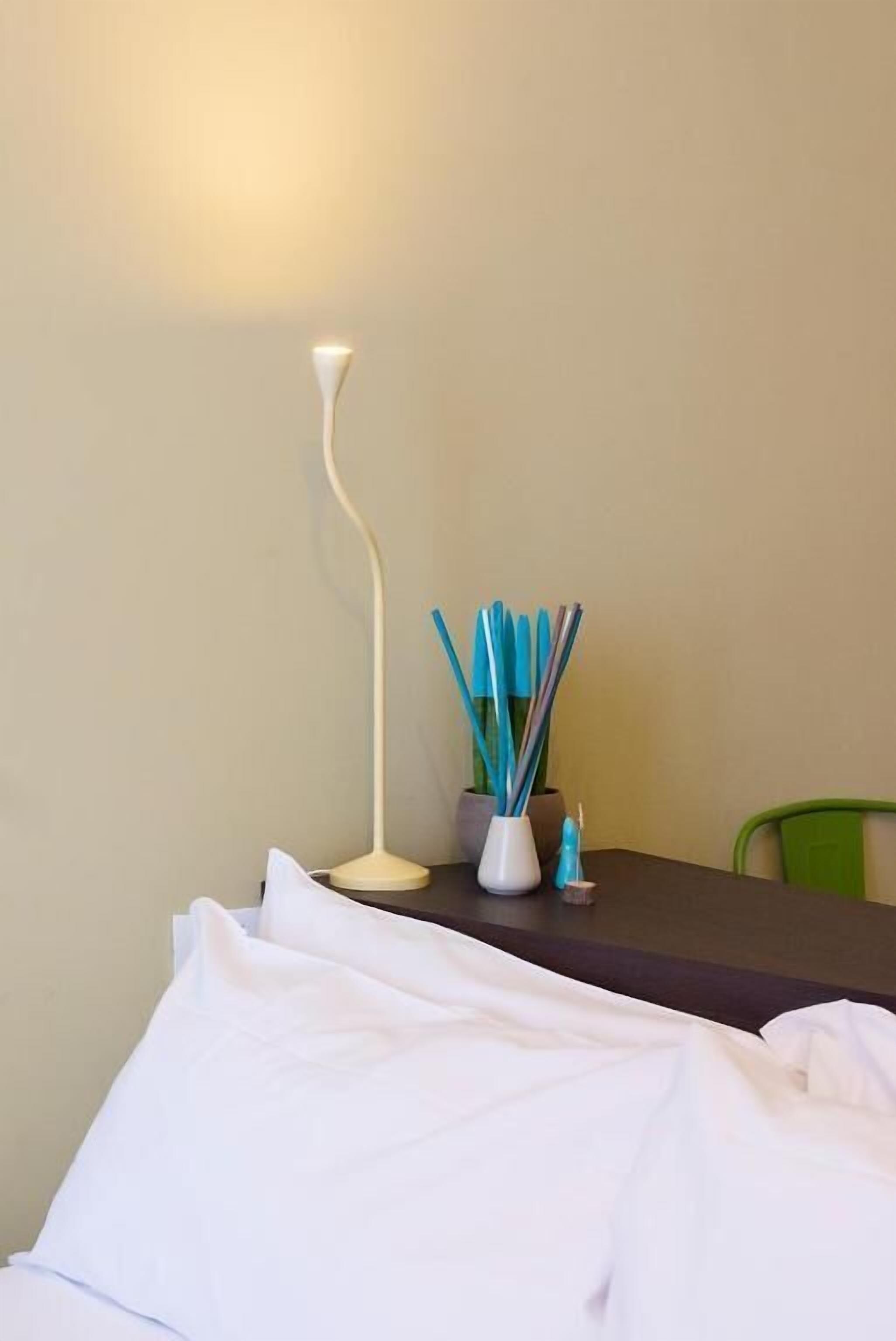 A bed with white bedding, a bedside table with a lamp and a vase of pencils, and a green chair in a room with plain walls.