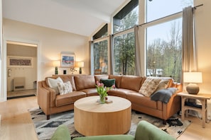 A living room with a brown leather sofa, a round coffee table with a vase of flowers, and large windows offering a view of trees. - Renovated Modern Chalet in Mad River Valley (Warren)