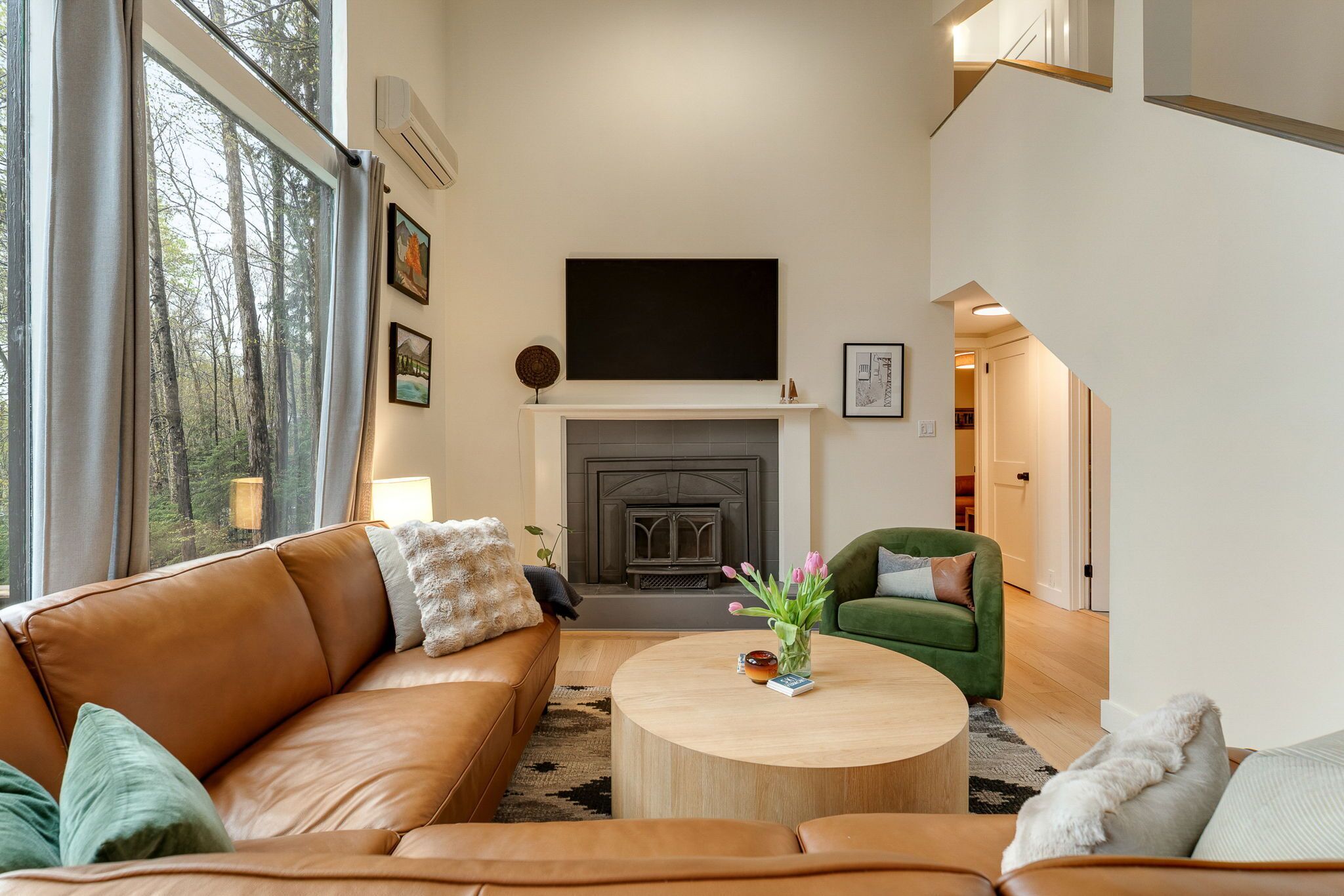 Een woonkamer met een open haard, een groene fauteuil, een bruine leren bank en een ronde salontafel met een bloevas.