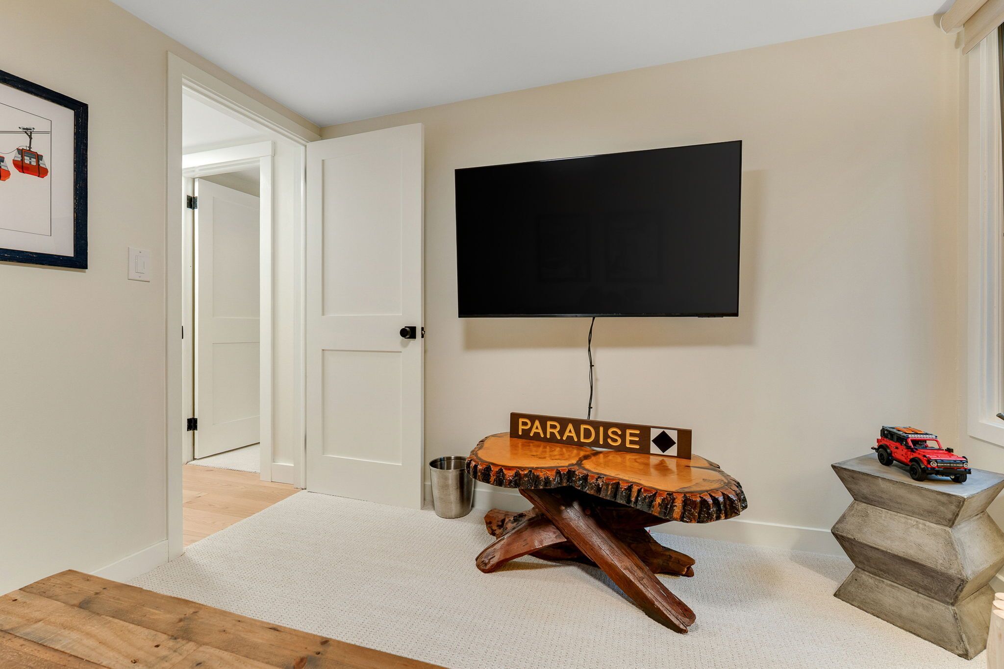 Een kamer met een wandtelevisie, een houten tafel met een ‘PARADISE’ bord, een speelgoedauto en een deur naar een andere kamer.