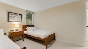 Un dormitorio con una cama individual, una mesita de noche con lámpara, un cuadro enmarcado en la pared y una ventana con vistas a árboles.