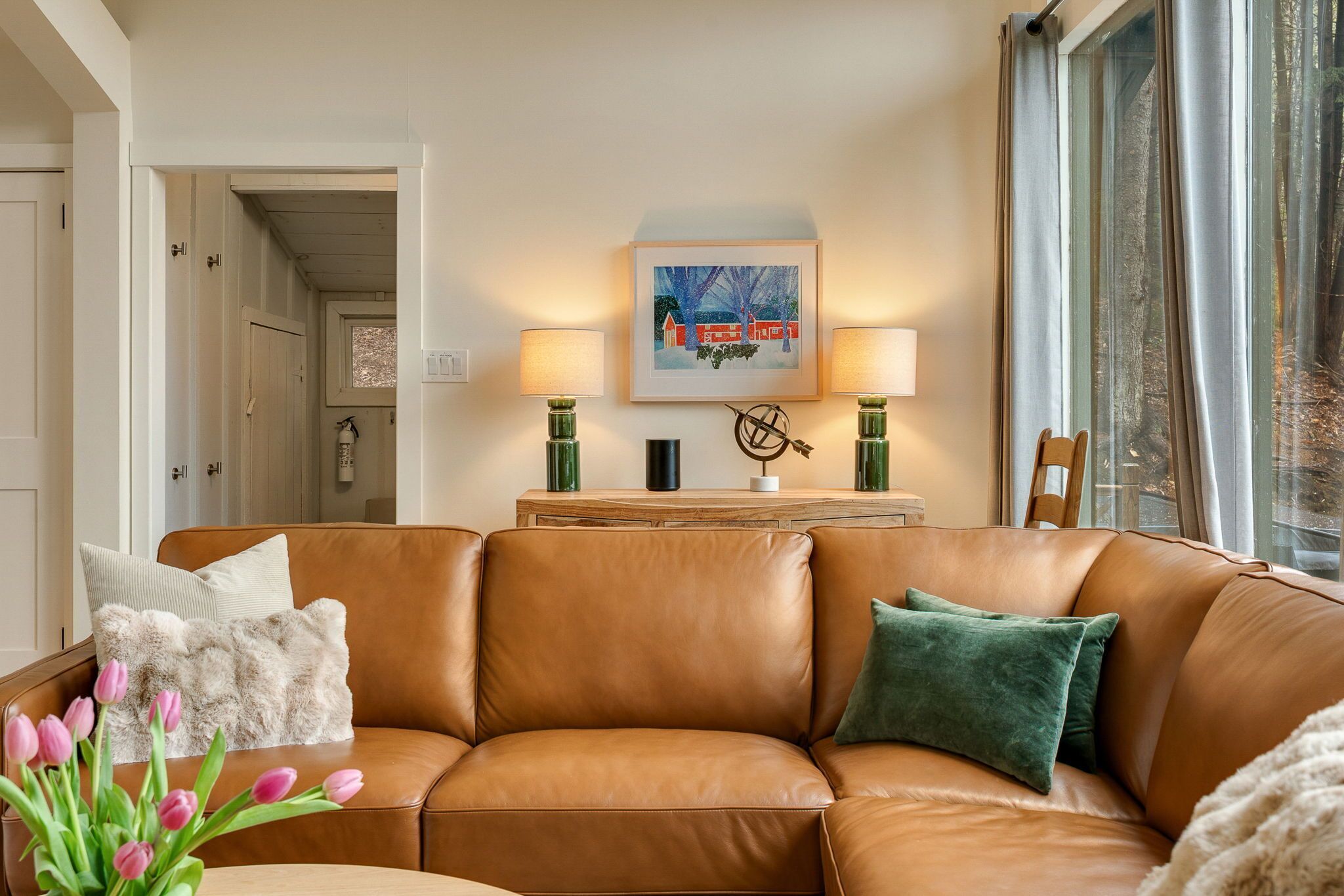 A brown leather sofa with a green pillow, a framed picture on the wall, and a window with curtains.