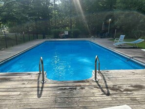 Una piscina rectangular con escaleras metálicas sobre una terraza de madera, rodeada por una valla y árboles.