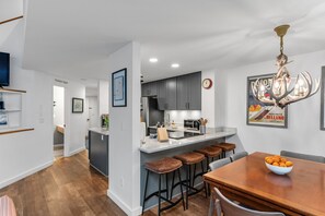 A modern kitchen with a central island, bar stools, and a dining table with a bowl of fruit. - Mad River Valley Modern 3 bedroom & 2 bath Condo; Convenient Location (Warren)