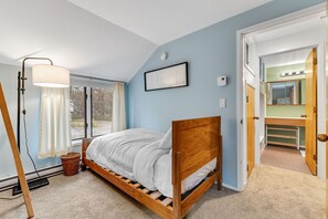 Un dormitorio con cama de madera, lámpara de pie, ventana con cortinas y puerta que da a un baño con espejo e iluminación.