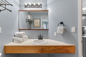 A bathroom with a wooden vanity, a white sink, a mirror with lights, a towel rack, and a framed picture on the wall. - Mad River Valley Modern 3 bedroom & 2 bath Condo; Convenient Location (Warren)