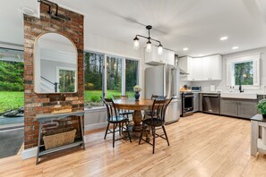 Une cuisine moderne avec un mur d’accent en briques, de grandes fenêtres et un coin repas comprenant une table ronde et des chaises.