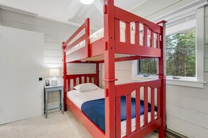 Un lit superposé avec un matelas blanc et une parure de lit bleue, une petite table de chevet avec une lampe et une fenêtre donnant sur des arbres.