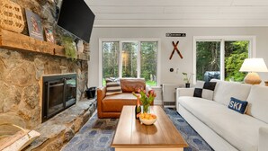 A cozy living room with a stone fireplace, a white couch, a brown leather chair, and a coffee table with a bottle and flowers. - The Warren Nook: Your cozy, modern mountain basecamp. (Warren)