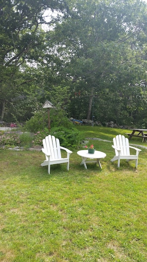Ein grünes Freigelände mit zwei weißen Adirondack-Stühlen und einem kleinen runden Tisch, umgeben von Bäumen und einem Picknicktisch im Hintergrund.