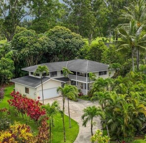 Una casa de dos pisos con balcón, rodeada de abundante follaje y palmeras.