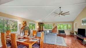 A spacious living area with a dining table set for four, a grey sofa, a television, and a ceiling fan. - Kanaloa Hale - Four Bedroom/Two and a Half Bath Home (Princeville)
