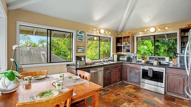 A spacious kitchen with a dining area, large windows, and a view of greenery outside. - Kanaloa Hale - Four Bedroom/Two and a Half Bath Home (Princeville)