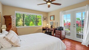 A hotel room with a bed, desk, chair, ceiling fan, window with blinds, and a view of greenery outside. - Kanaloa Hale - Four Bedroom/Two and a Half Bath Home (Princeville)
