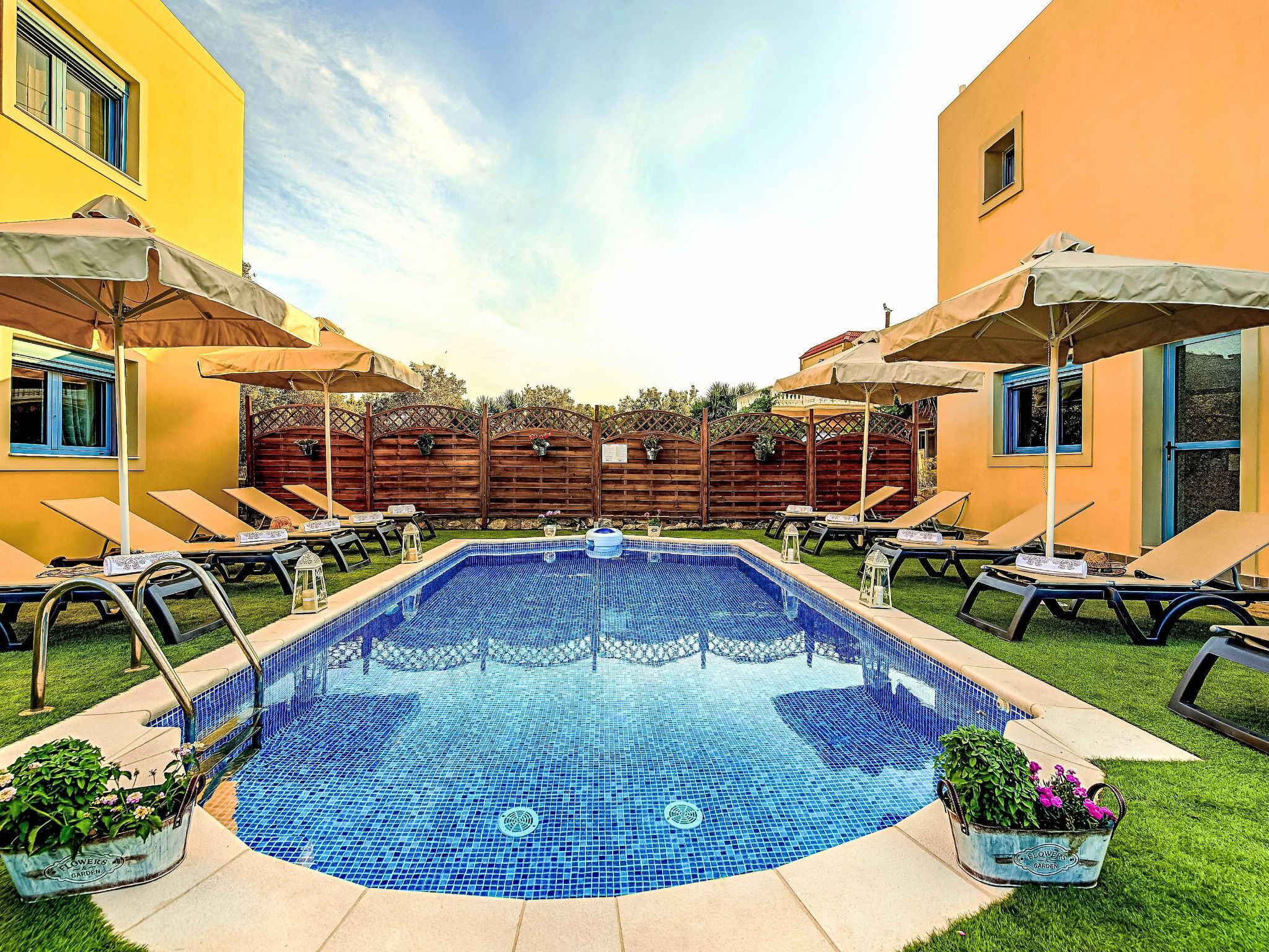 A swimming pool with lounge chairs and umbrellas, surrounded by a wooden fence and a yellow building.