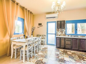A dining area with a table, chairs, a vase with dried flowers, a chandelier, and a kitchen with patterned tiles and cabinets. - Retro Villas - Superior with Pool by Interhome (Pefki)
