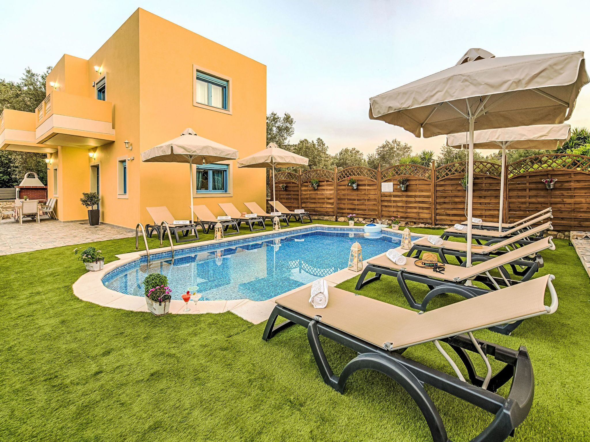 A swimming pool with lounge chairs and umbrellas, surrounded by a wooden fence and a building with a yellow facade.