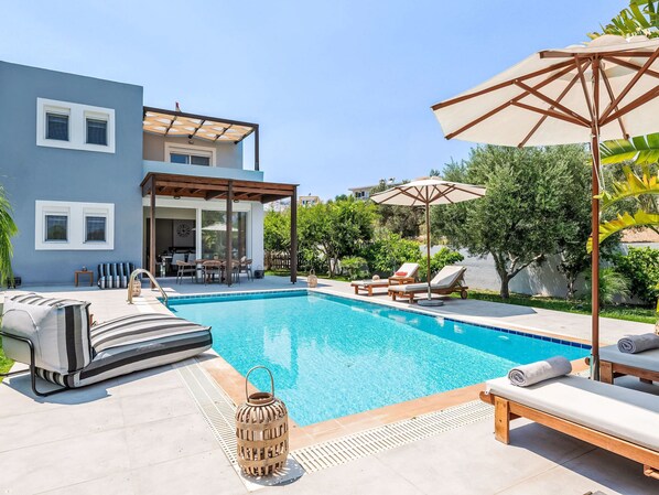 Una piscina con sillones de descanso y sombrillas, un patio cubierto y una casa moderna con amplias ventanas.
