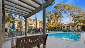 A wooden swing bench under a pergola, a swimming pool, and a poolside lounge area with sun loungers and umbrellas. - 
Coastal Retreat Steps from Coligny Beach, Dining, Shops, and Celebration Park! (Hilton Head Island)