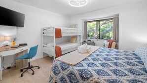 A room with bunk beds, a desk, a chair, a bed with a patterned bedspread, a television, and a window with a view of trees. - 
Coastal Retreat Steps from Coligny Beach, Dining, Shops, and Celebration Park! (Hilton Head Island)