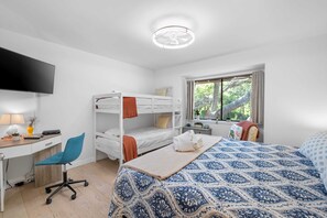 A room with bunk beds, a desk, a chair, a bed with a patterned bedspread, a television, and a window with a view of trees. - 
Coastal Retreat Steps from Coligny Beach, Dining, Shops, and Celebration Park! (Hilton Head Island)