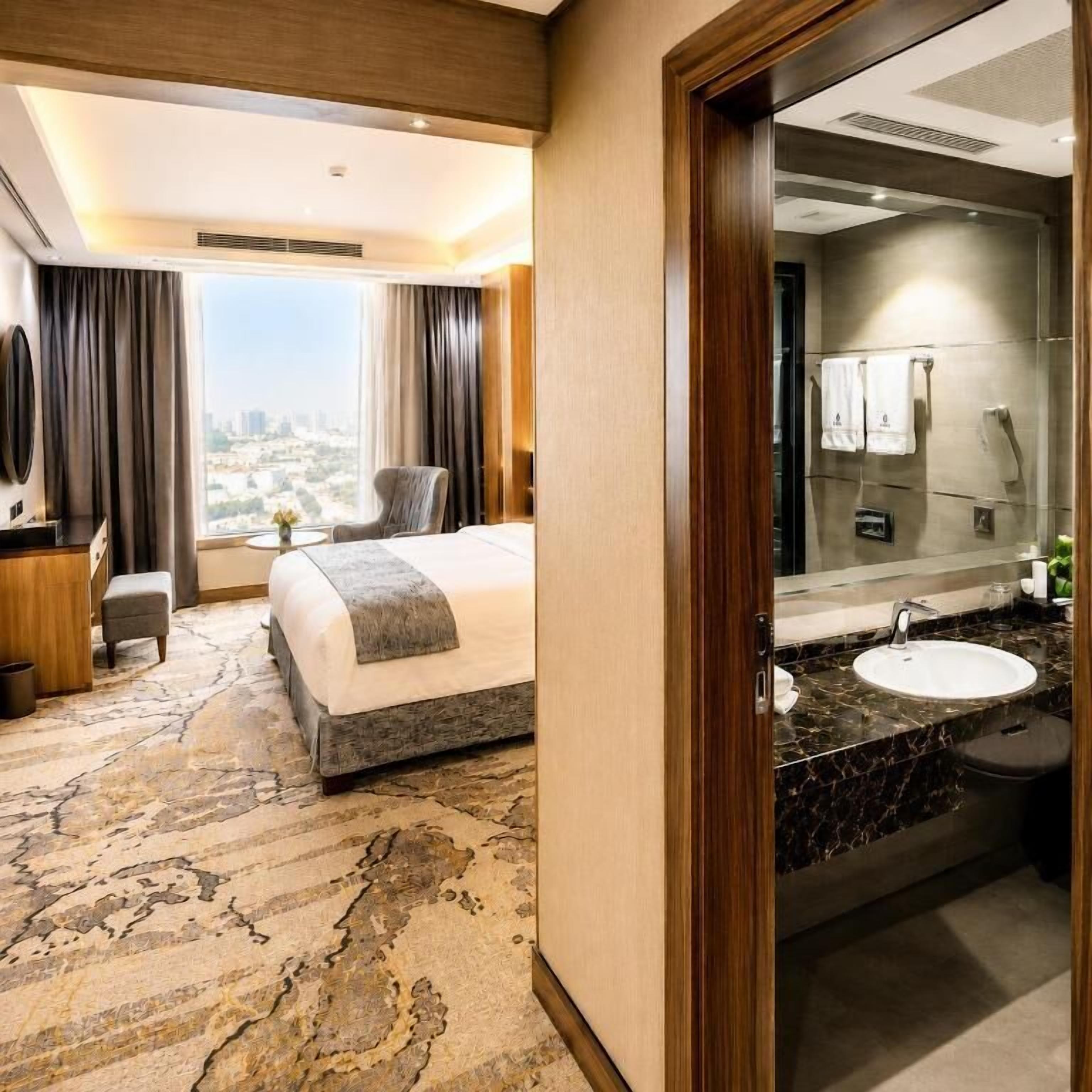 Habitación de hotel con cama, escritorio, silla, baño con lavamanos y espejo, y vista a la ciudad.