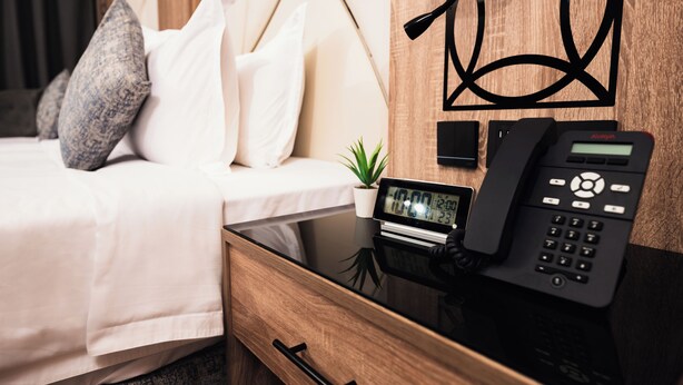 A hotel room with a bed, a bedside table with a phone, a small clock, and a drawer. - Medhal Hotel (Jeddah)
