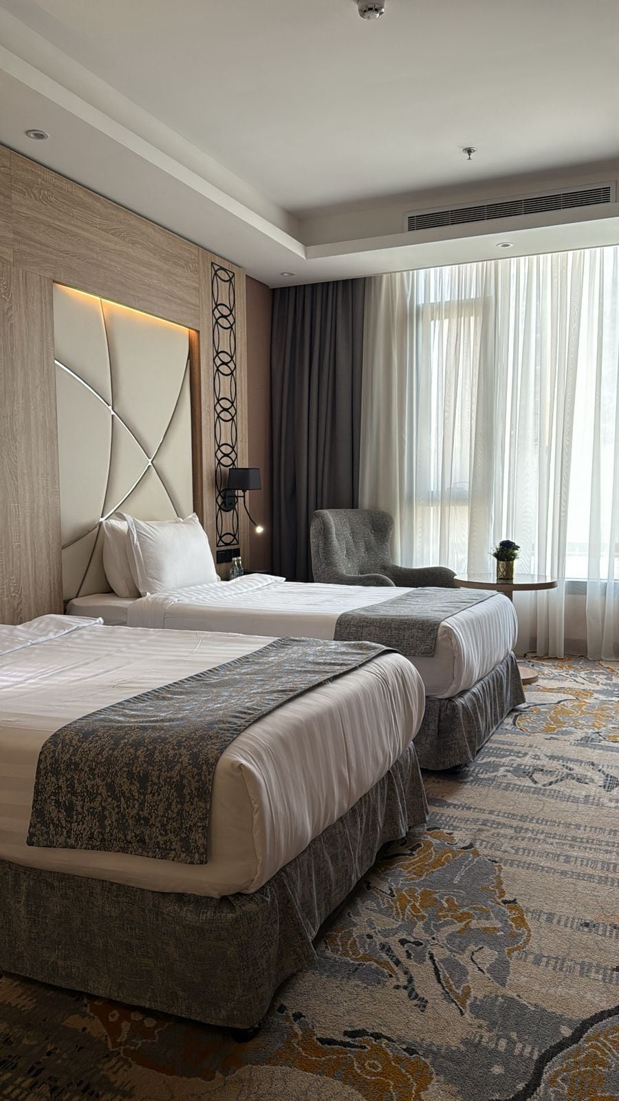 A hotel room with two beds, a headboard with a geometric design, a small table with a lamp, and a large window with sheer curtains.