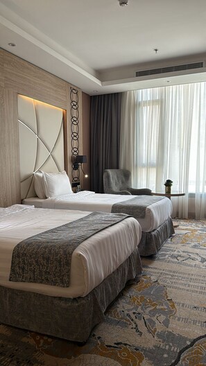 A hotel room with two beds, a headboard with a geometric design, a small table with a lamp, and a large window with sheer curtains. - Medhal Hotel (Jeddah)