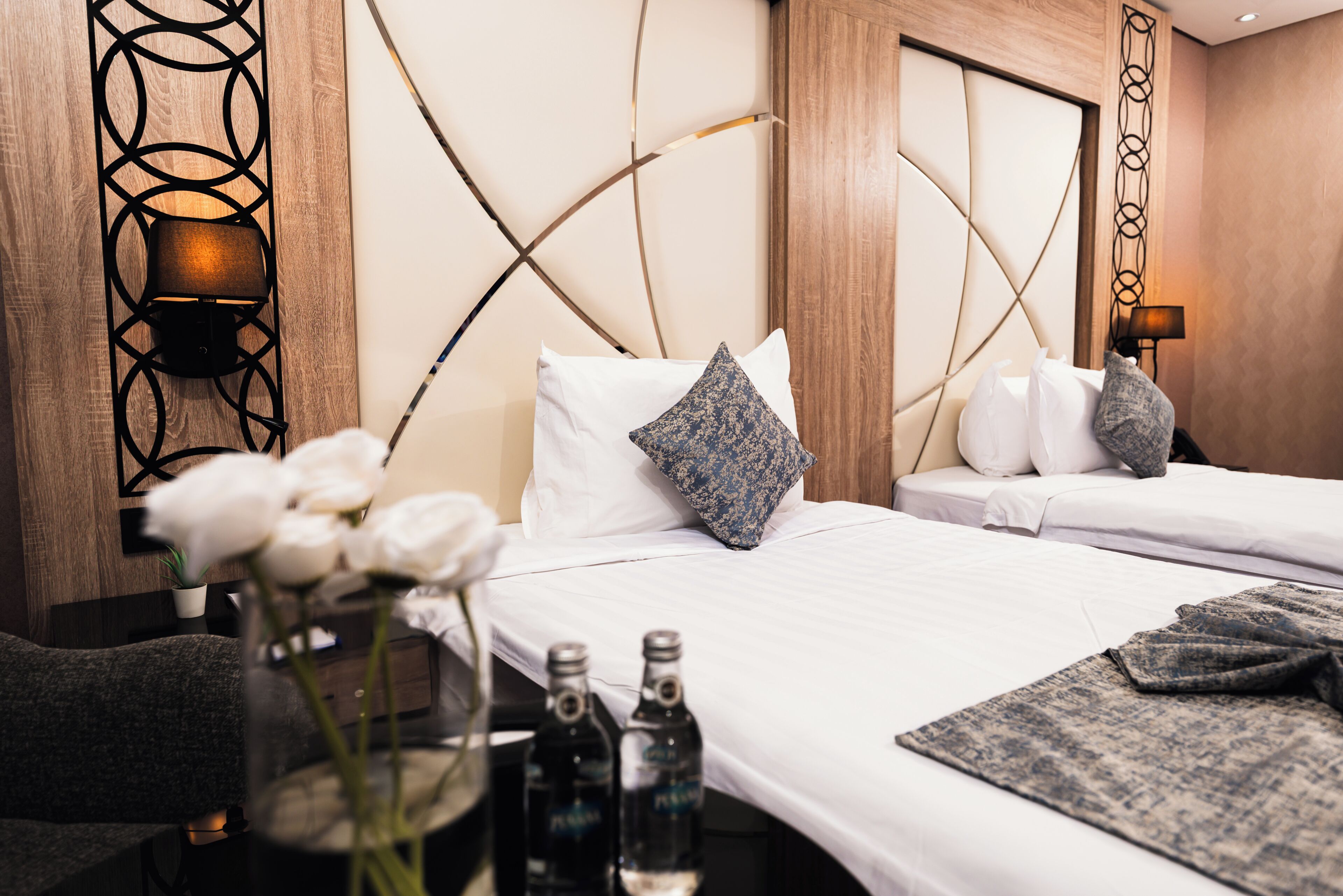 Habitación de hotel con una cama, dos almohadas, una mesita de noche, un florero con flores y dos botellas de agua.